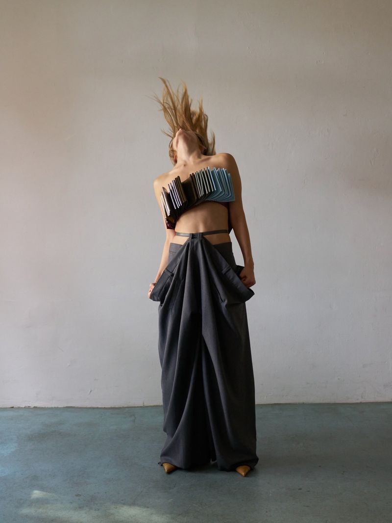 Adult female model in a studio wearing an avant-garde blue and black top and a long gray draped skirt, tossing her blond hair.