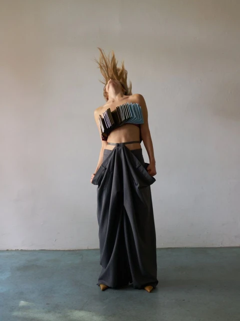 Adult female model in a studio wearing an avant-garde blue and black top and a long gray draped skirt, tossing her blond hair.