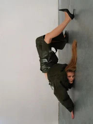 A model in an olive green leather outfit and black stiletto heels poses in an inverted contorted position on a studio floor.