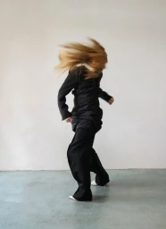 Model with blurred whipping blonde hair wearing an oversized black tailored suit and white pointed shoes against a minimal white studio wall.