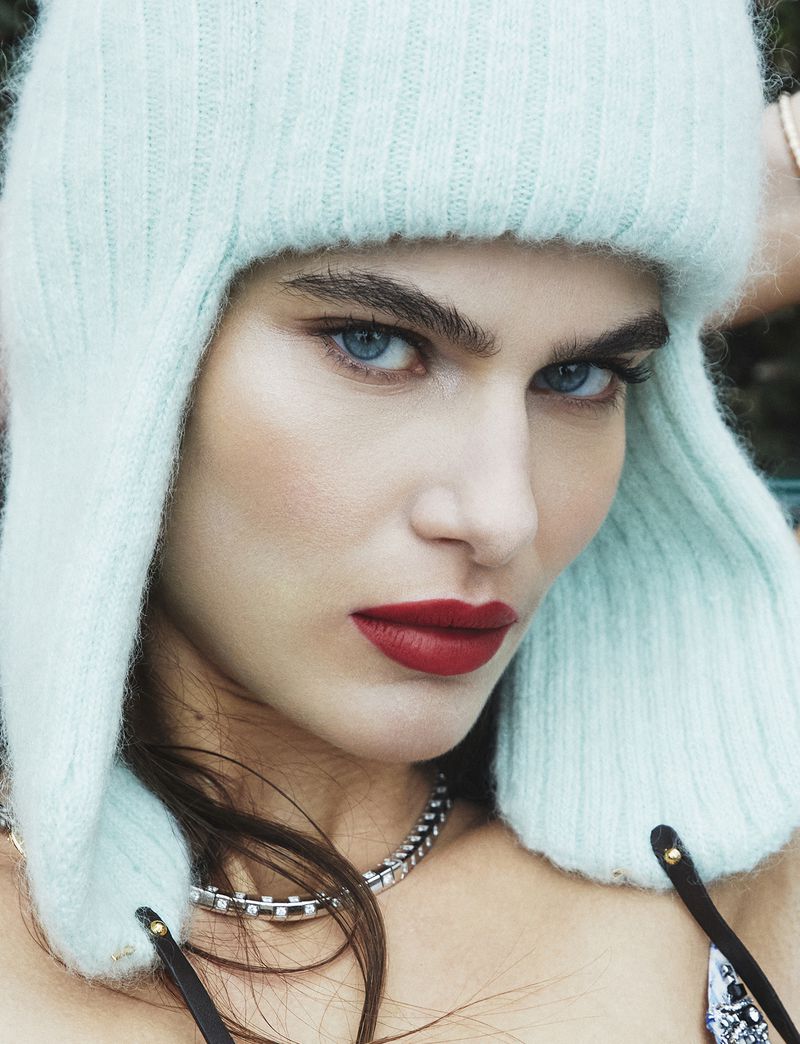 Close-up portrait of a woman with blue eyes and red lipstick, wearing a light-blue knitted trapper hat. Fashion beauty shot.