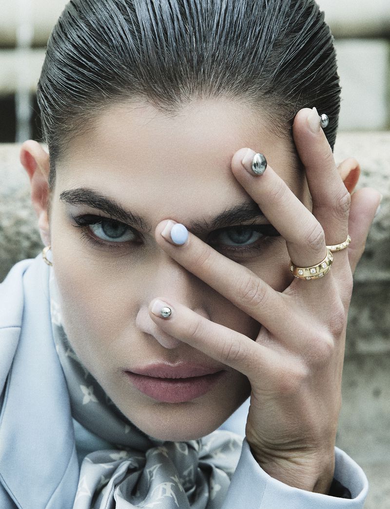 Close-up of a brunette adult female fashion model with light skin and blue eyes, wearing pastel makeup, and a light blue jacket.