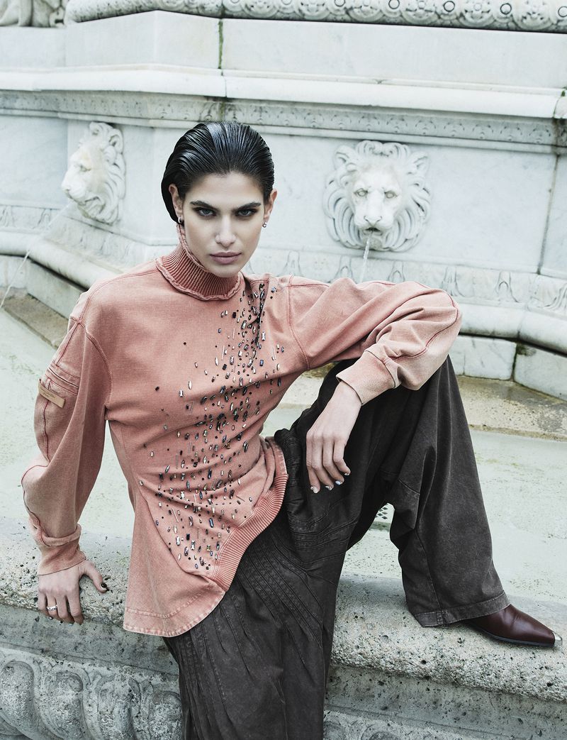 A woman in an orange top and dark pants sits on a stone structure, with a fountain featuring lion sculptures in the background.