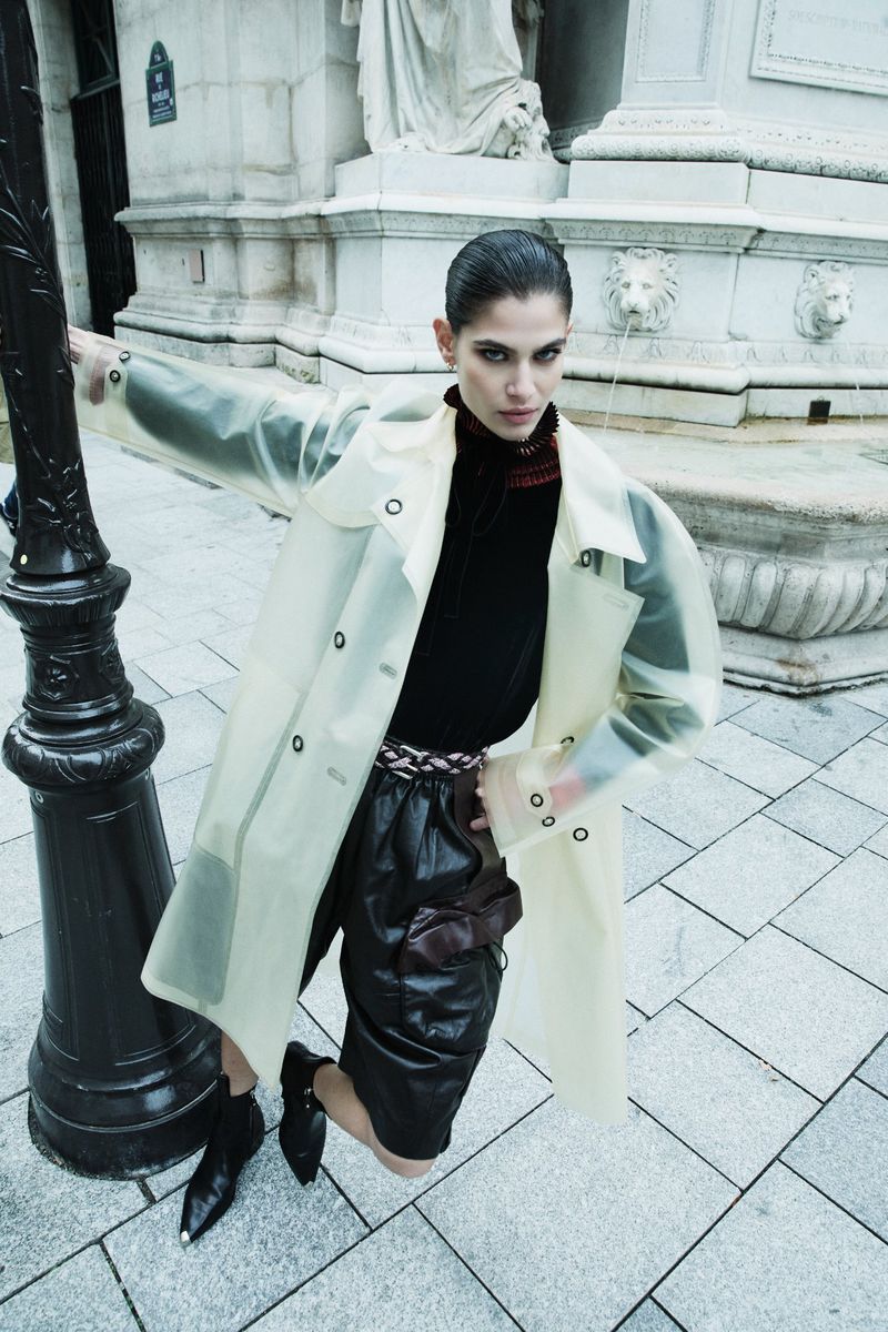 Fashionable adult woman in a translucent trench coat, black top and shorts poses confidently in an urban setting by a black lamp post.