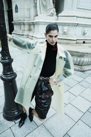 Fashionable adult woman in a translucent trench coat, black top and shorts poses confidently in an urban setting by a black lamp post.