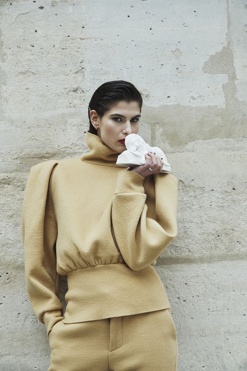 A woman in a yellow turtleneck outfit holds a white sculpture against a textured concrete wall in a medium shot fashion editorial.