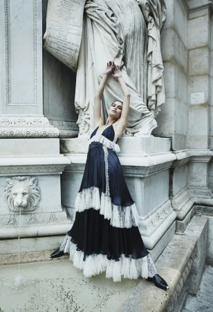 Fashionable woman in a long, tiered dress posing in front of a classical stone fountain with lion head sculptures, outdoors.