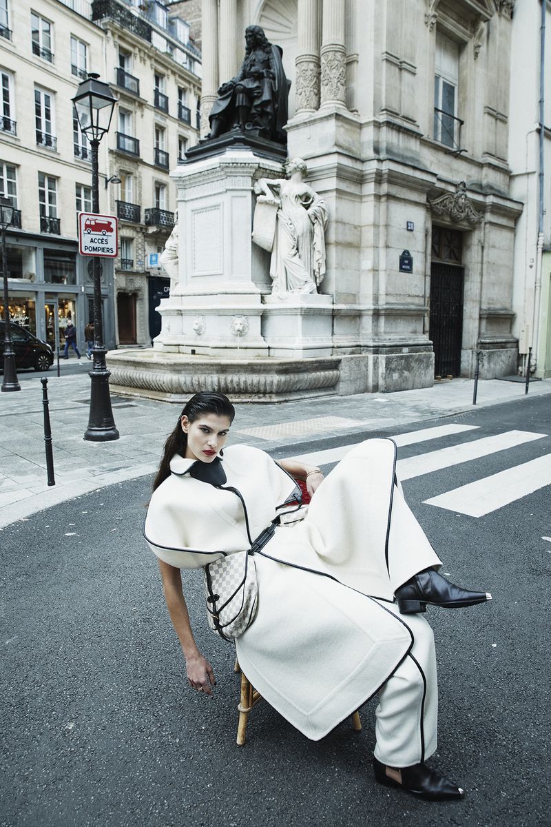 Fashion shot of a woman in a white coat and pants sitting on a chair in a city street with a monument and buildings in the background.