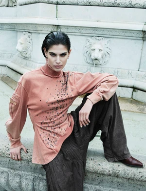 Model Anthi Fakidari in a peach embellished top and dark trousers posed against a carved stone fountain structure