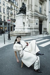 Model Anthi Fakidari in an oversized cream cape coat and trousers seated in a chair on an asphalt street in front of a monument.