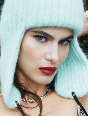 Close-up portrait of Anthi Fakidari wearing a mint green knitted ushanka hat with bold red lipstick and a silver chain necklace.