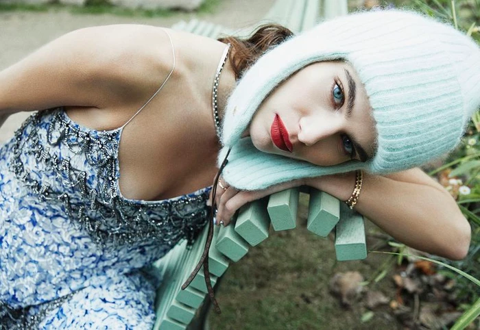 Model Anthi Fakidari rests on a light green bench wearing a mint knit trapper hat and blue floral beaded slip dress.