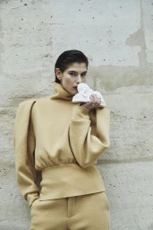 Anthi Fakidari wearing a structured beige high-neck top and holding a miniature white sculpture against a stone wall.
