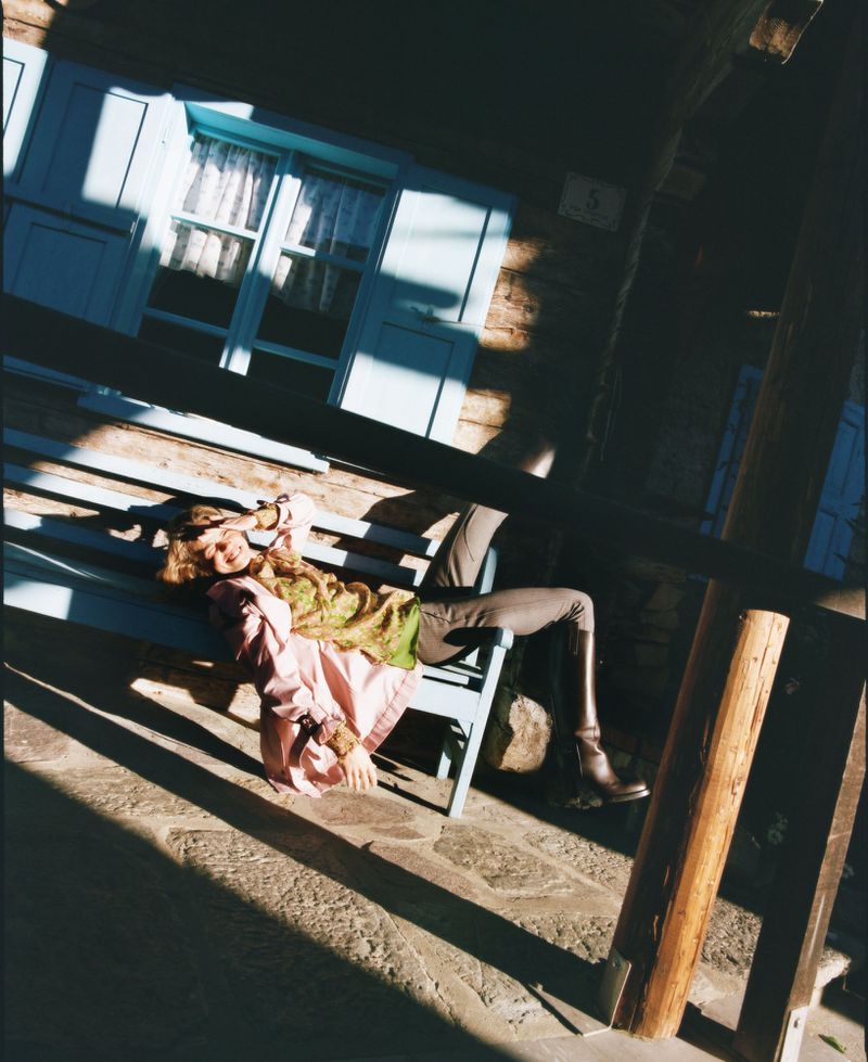 A woman relaxes on a blue bench outside a weathered building, dappled in sunlight, wearing olive, brown, pink, and boots.