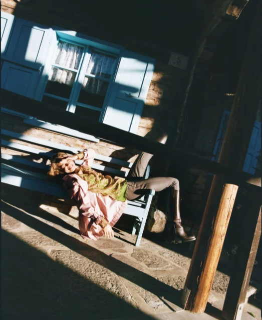 A woman relaxes on a blue bench outside a weathered building, dappled in sunlight, wearing olive, brown, pink, and boots.