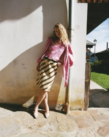 Woman in pink blouse and striped skirt leans against wall. Outdoor fashion shot with natural light, neutral tones and subtle shadows.