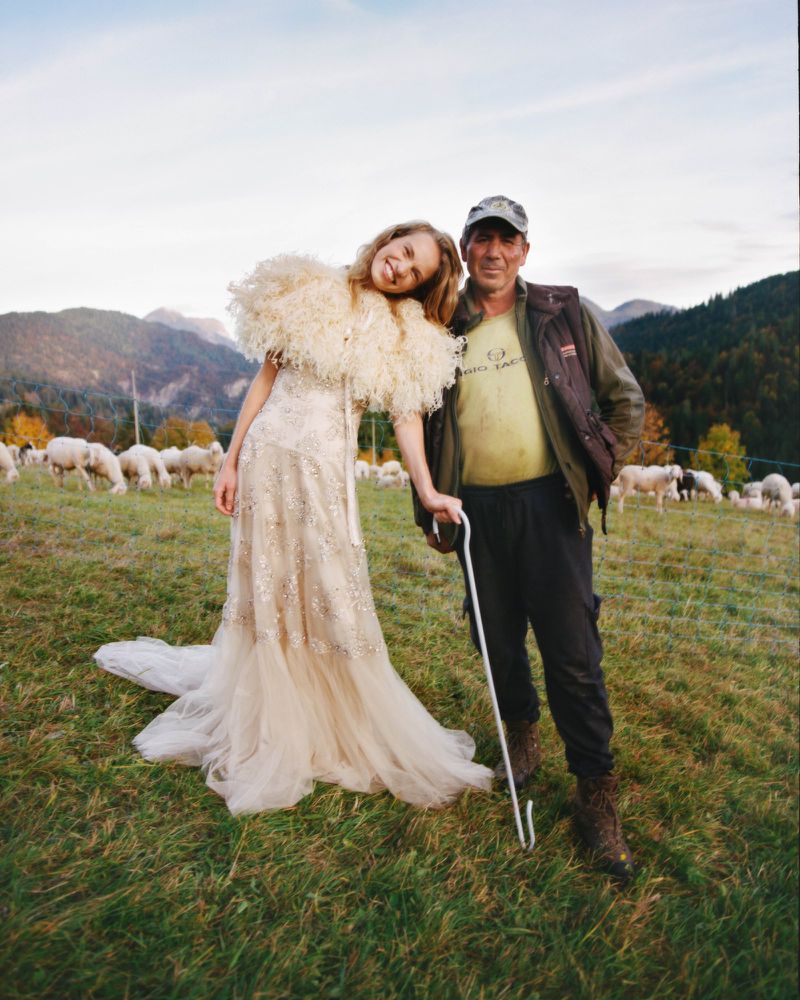 Woman in gown poses with shepherd in mountain field with grazing sheep, creating a surreal fashion editorial moment in nature.