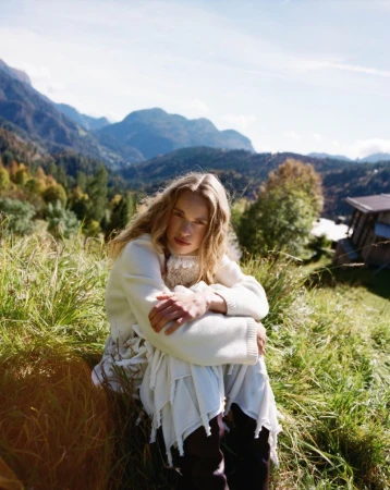 Woman with long blonde hair wearing a white sweater and skirt sits in tall grass against mountain backdrop in a scenic outdoor setting.