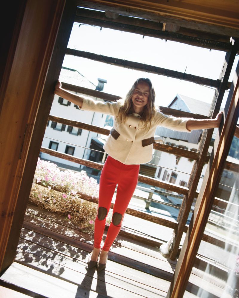 A cheerful young woman in red pants and a white jacket stands in an open doorway, smiling with arms outstretched, enjoying a bright day.