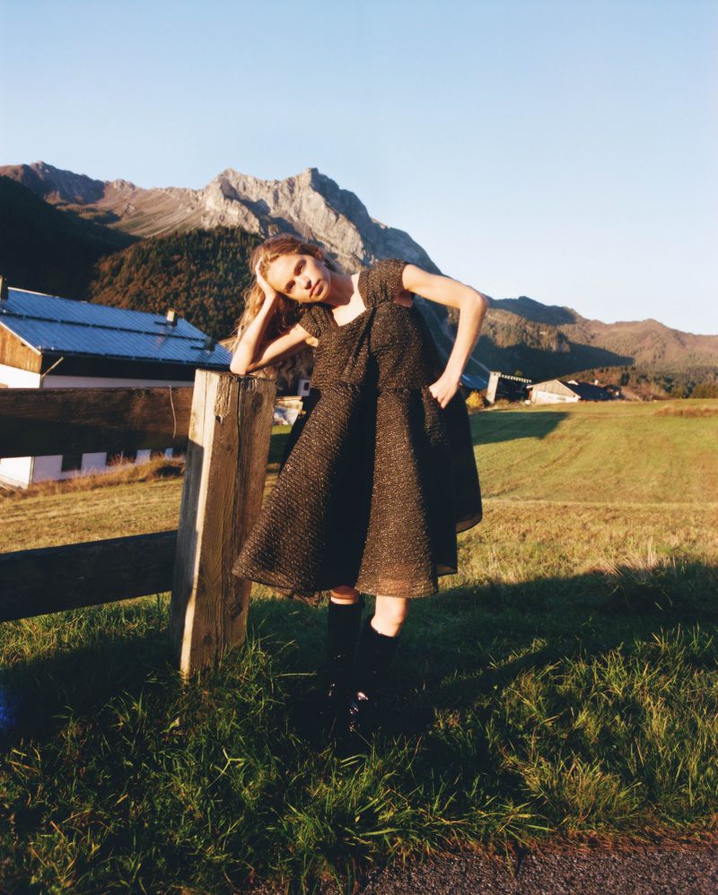 A fair-skinned woman in a brown dress stands beside a wooden fence in a grassy field against a backdrop of mountains and a clear sky.