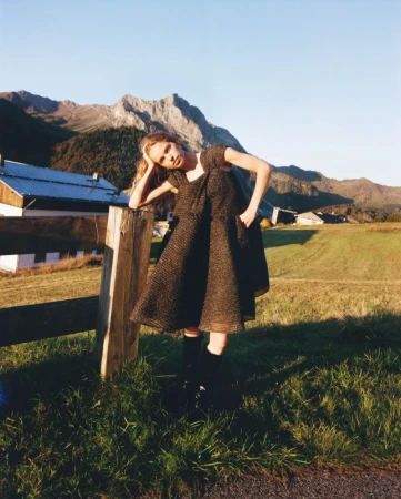 A fair-skinned woman in a brown dress stands beside a wooden fence in a grassy field against a backdrop of mountains and a clear sky.