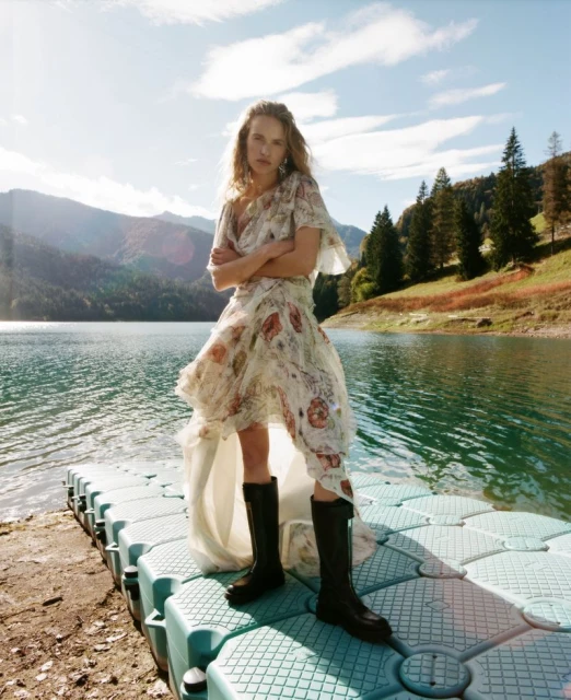 Woman in floral dress and boots poses on floating dock with lake, mountain, and sky background on a sunny day. Fashion editorial shoot.