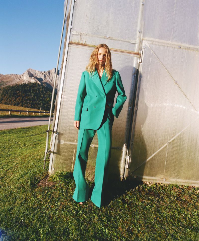 An adult female fashion model poses in a green suit in front of a building on a sunny day, with mountains in the background.