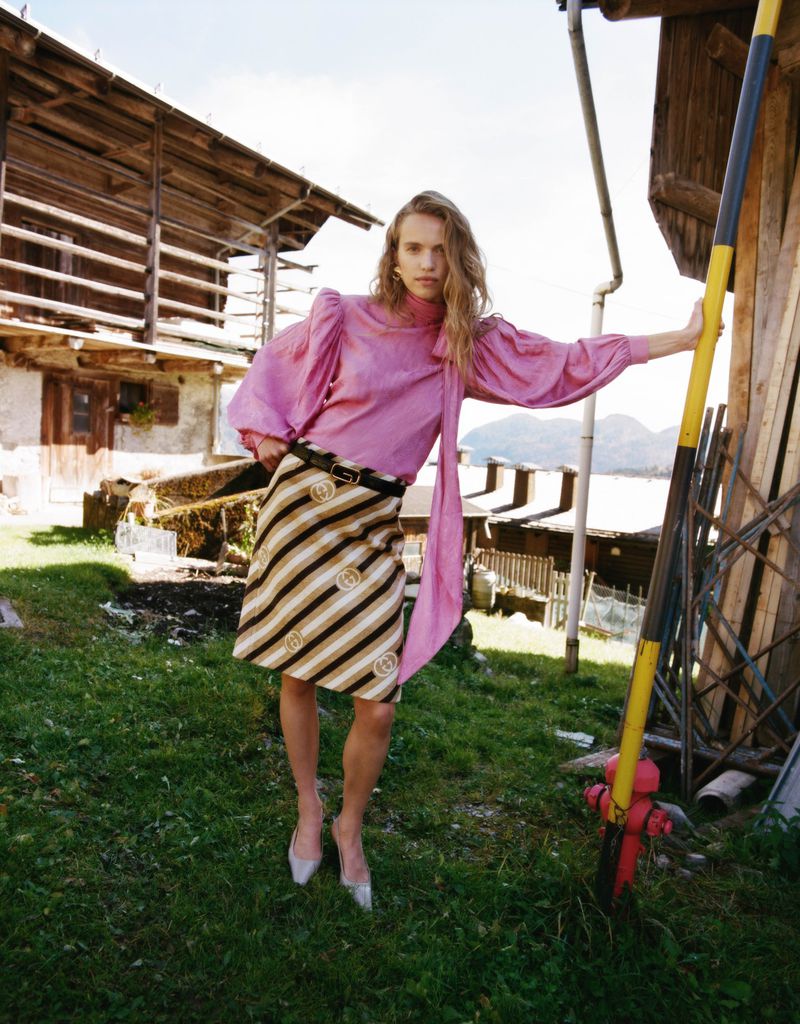 A fashionable woman in a pink blouse and striped skirt poses outdoors in front of a rural building and green grass.