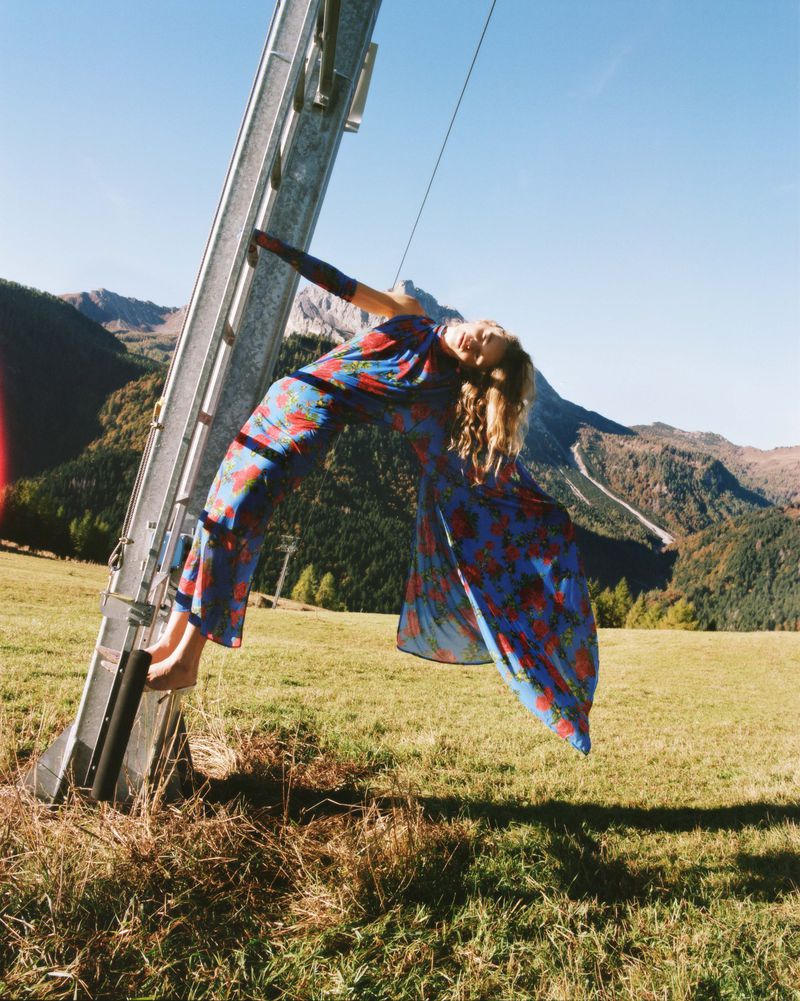 A woman in a floral dress poses on a metal structure in a grassy field with mountains in the background on a sunny day.