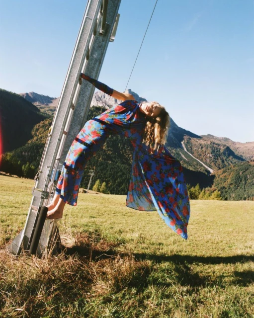 A woman in a floral dress poses on a metal structure in a grassy field with mountains in the background on a sunny day.