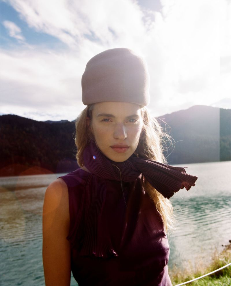 Portrait of a blonde woman wearing a hat and scarf, posing outdoors with lake and mountains in the background under bright, sunny conditions.