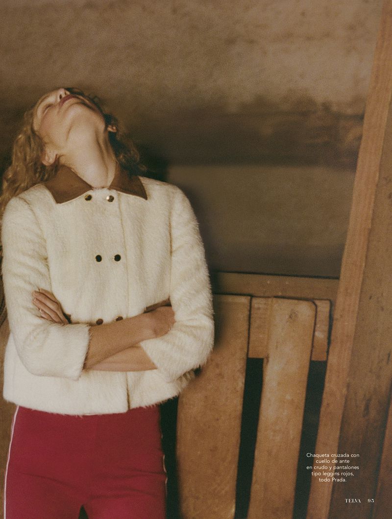 Woman in a fuzzy off-white jacket with a brown collar and bright pink pants poses indoors, illuminated by soft, warm lighting.