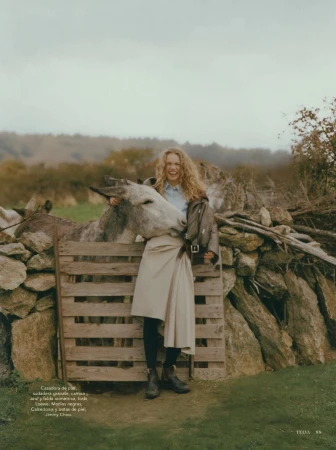 Fashion editorial image featuring a woman in a skirt, leather jacket, and boots, next to a donkey by a wooden gate in a rural setting.