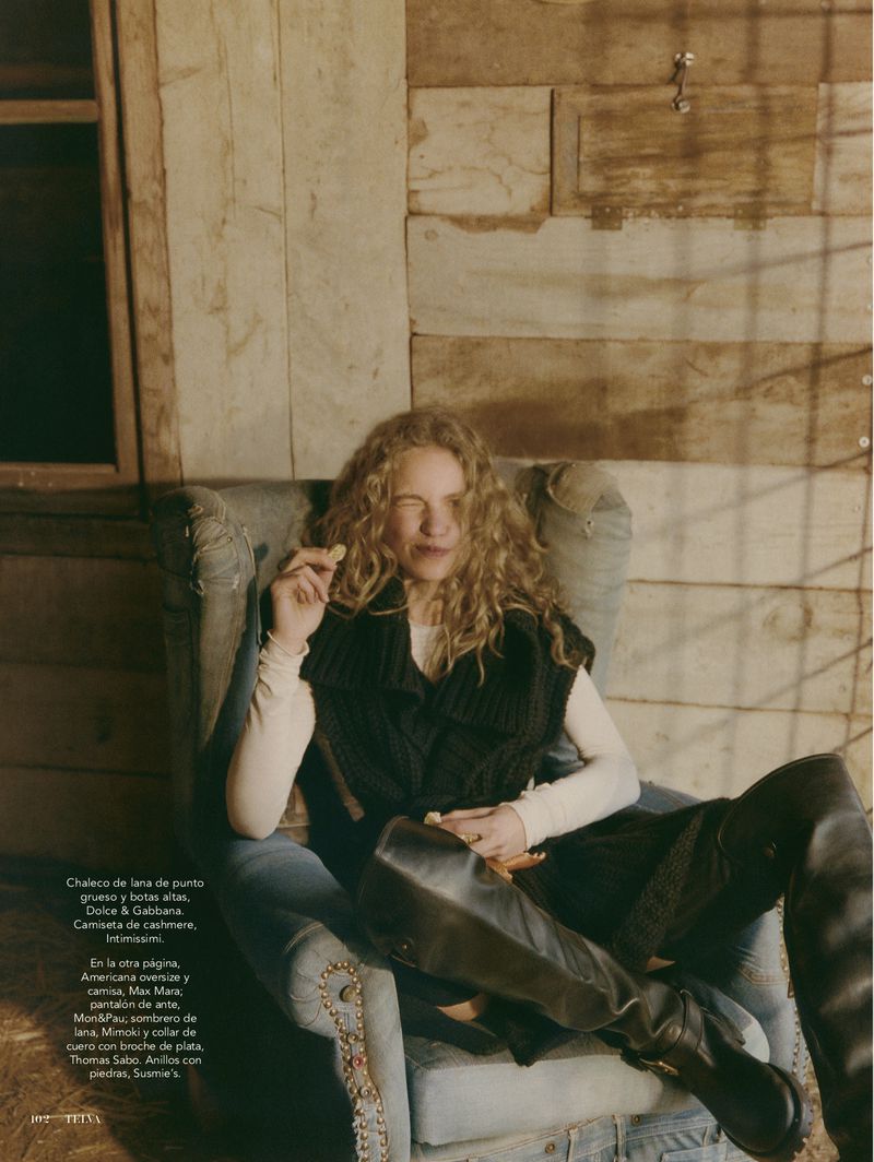 Fashion editorial shot of a woman in a chair wearing a black sweater vest and black over-the-knee boots in a rustic, wood setting.
