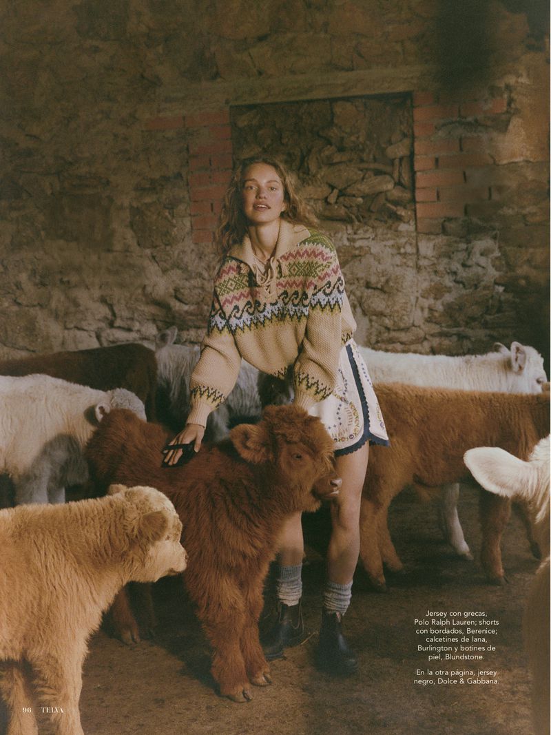 Editorial fashion shot of a young adult woman interacting with a group of tan and brown Highland calves inside of a stone barn.