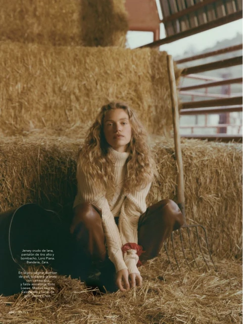 Fashion editorial photo of a woman with curly hair in a sweater and brown pants sitting on hay bales in a rustic barn.