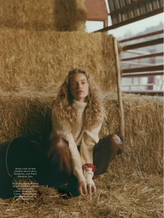 Fashion editorial photo of a woman with curly hair in a sweater and brown pants sitting on hay bales in a rustic barn.