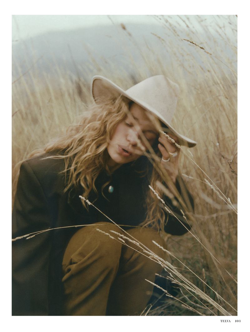 Woman with curly hair and a cowboy hat crouches in a field of tall brown grasses, wearing a blazer and brown pants in outdoor setting.