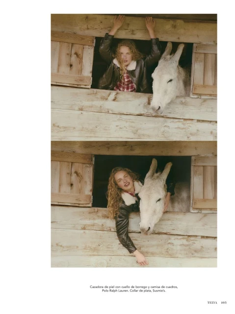 A woman with curly blonde hair wearing a brown jacket and plaid shirt poses in a barn window next to a white donkey.