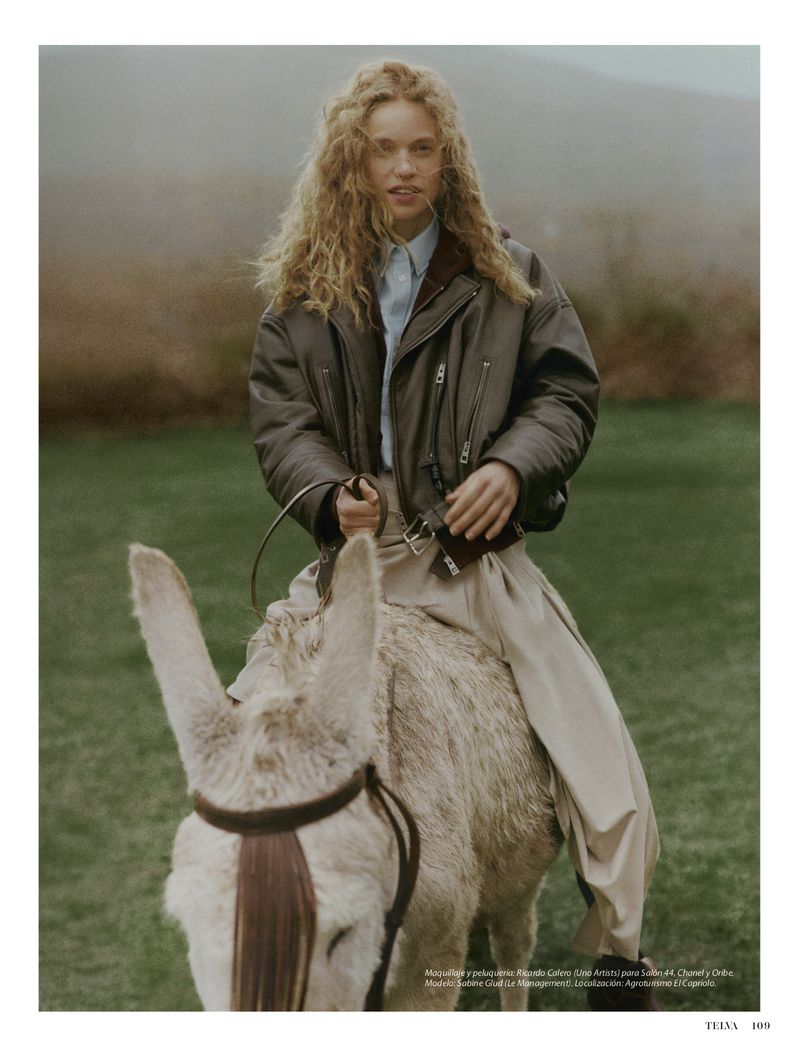 Woman in jacket riding a donkey in a grassy, outdoor setting. The donkey is white, and a grassy hill with sparse trees appears in the background.