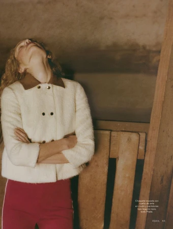 Sabine Glud in a cream fuzzy jacket and red trousers with arms crossed and head tilted back against a rustic wooden interior.