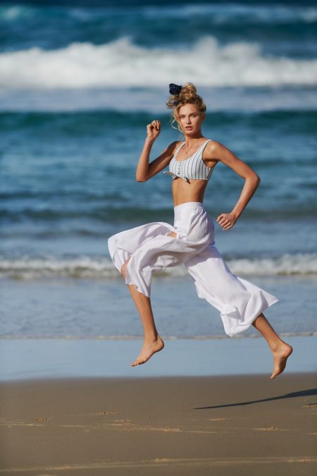 A young woman leaps mid-air on a sandy beach, sporting a bikini top and loose white pants, with a backdrop of ocean waves and sky.