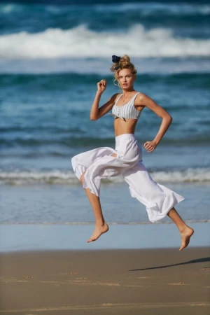 A young woman leaps mid-air on a sandy beach, sporting a bikini top and loose white pants, with a backdrop of ocean waves and sky.