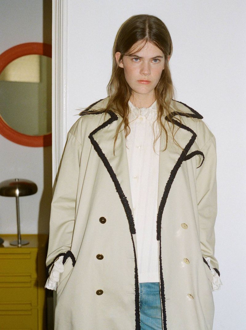 Indoor portrait of a woman wearing a trench coat and a frilly white blouse, posing in front of a white wall with decor.