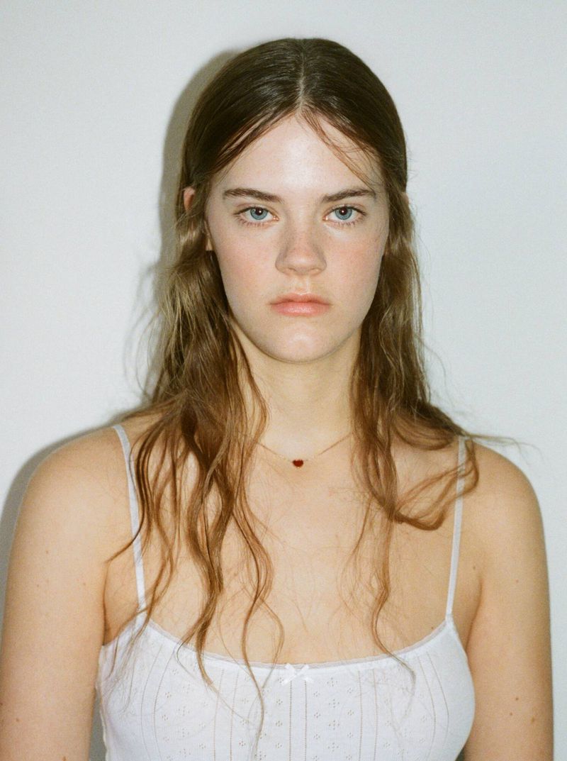 Portrait of a young Caucasian woman with long brown hair wearing a white floral-patterned tank top against a white background.