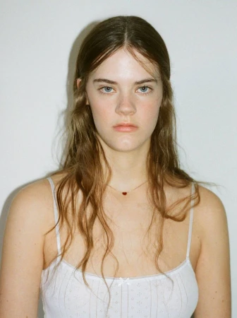 Portrait of a young Caucasian woman with long brown hair wearing a white floral-patterned tank top against a white background.