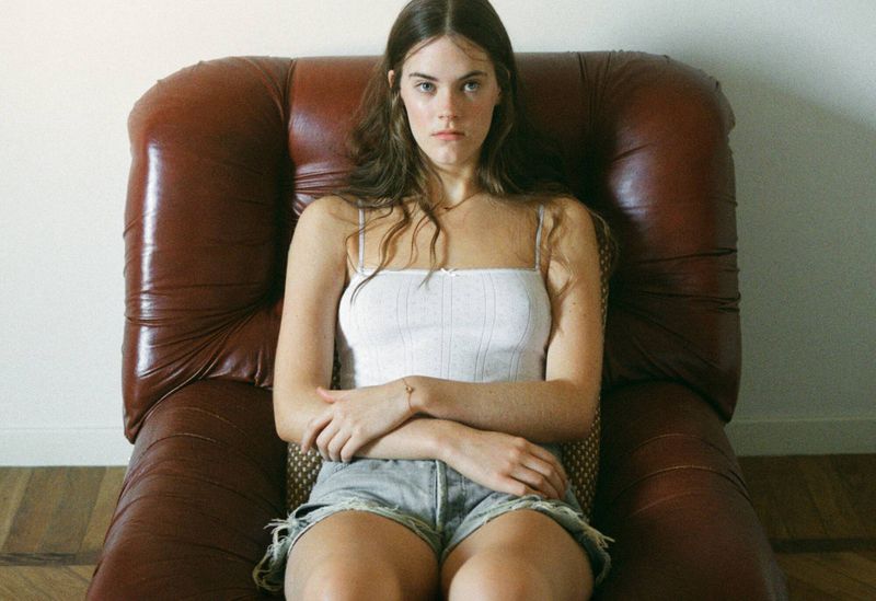 Young woman with long hair relaxes in an armchair wearing a white top and jean shorts in an indoor setting with neutral lighting.