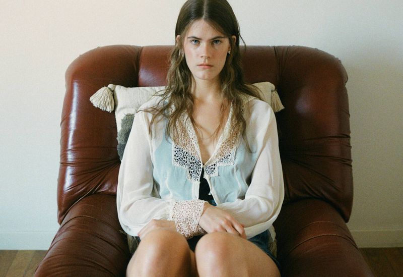 Young woman with long brown hair sits in a brown leather armchair indoors near a white wall, wearing a white top with lace details.
