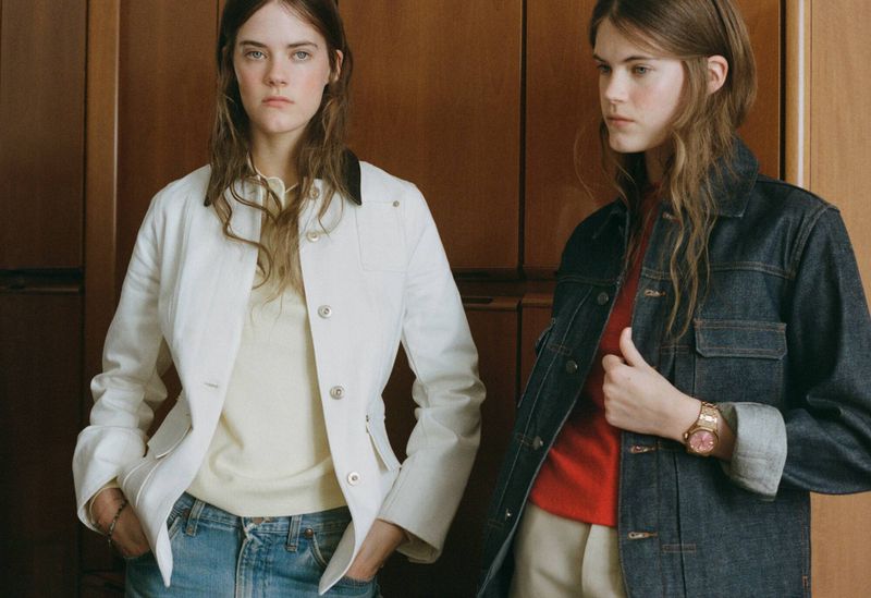 Two women in denim jackets pose indoors against a wood panel backdrop, exuding a casual yet stylish fashion vibe with natural lighting.