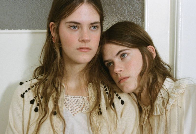 Portrait of two young women with long brown hair, pale skin, and light eyes, posing indoors in light-colored vintage-style clothing.
